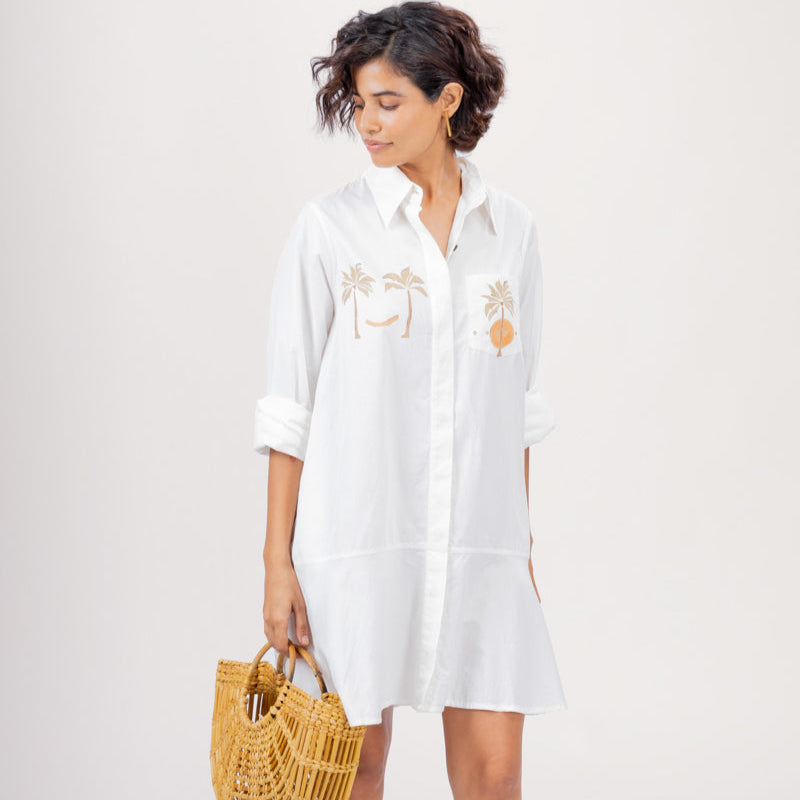Woman wearing a white shirt dress with floral embroidery, holding a woven bag on a plain background
