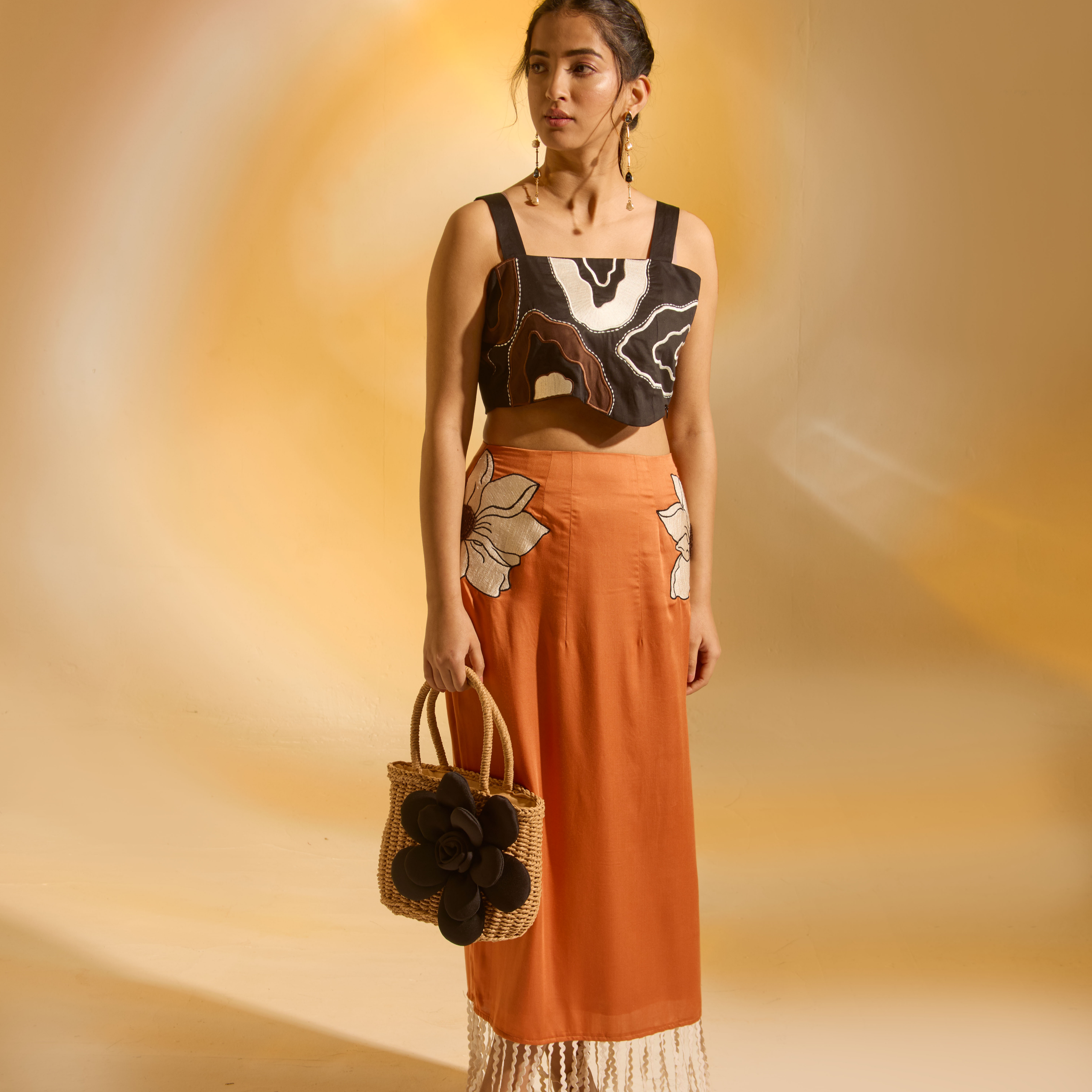 Woman in a black and white top with floral design and an orange skirt holding a bag against a warm-toned background