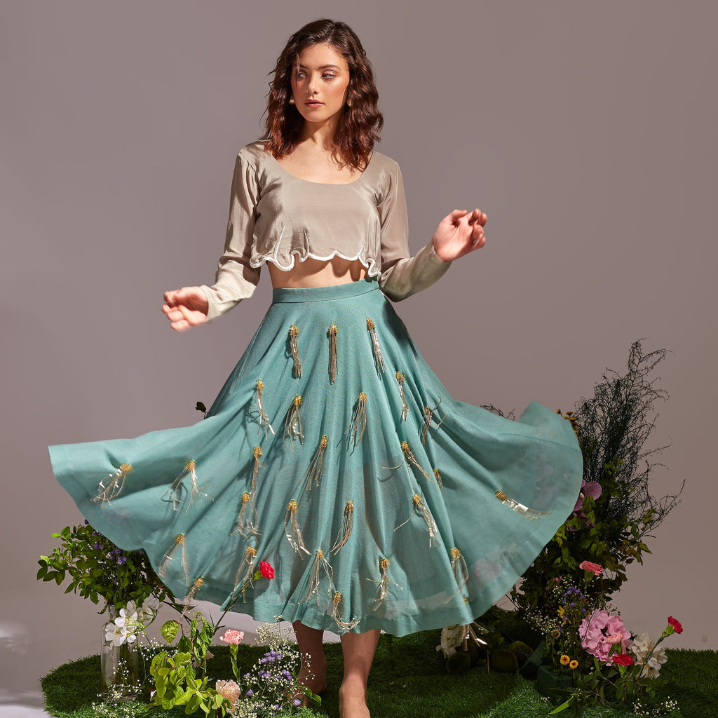 Woman in a teal skirt with floral embroidery standing on a grassy patch with flowers.