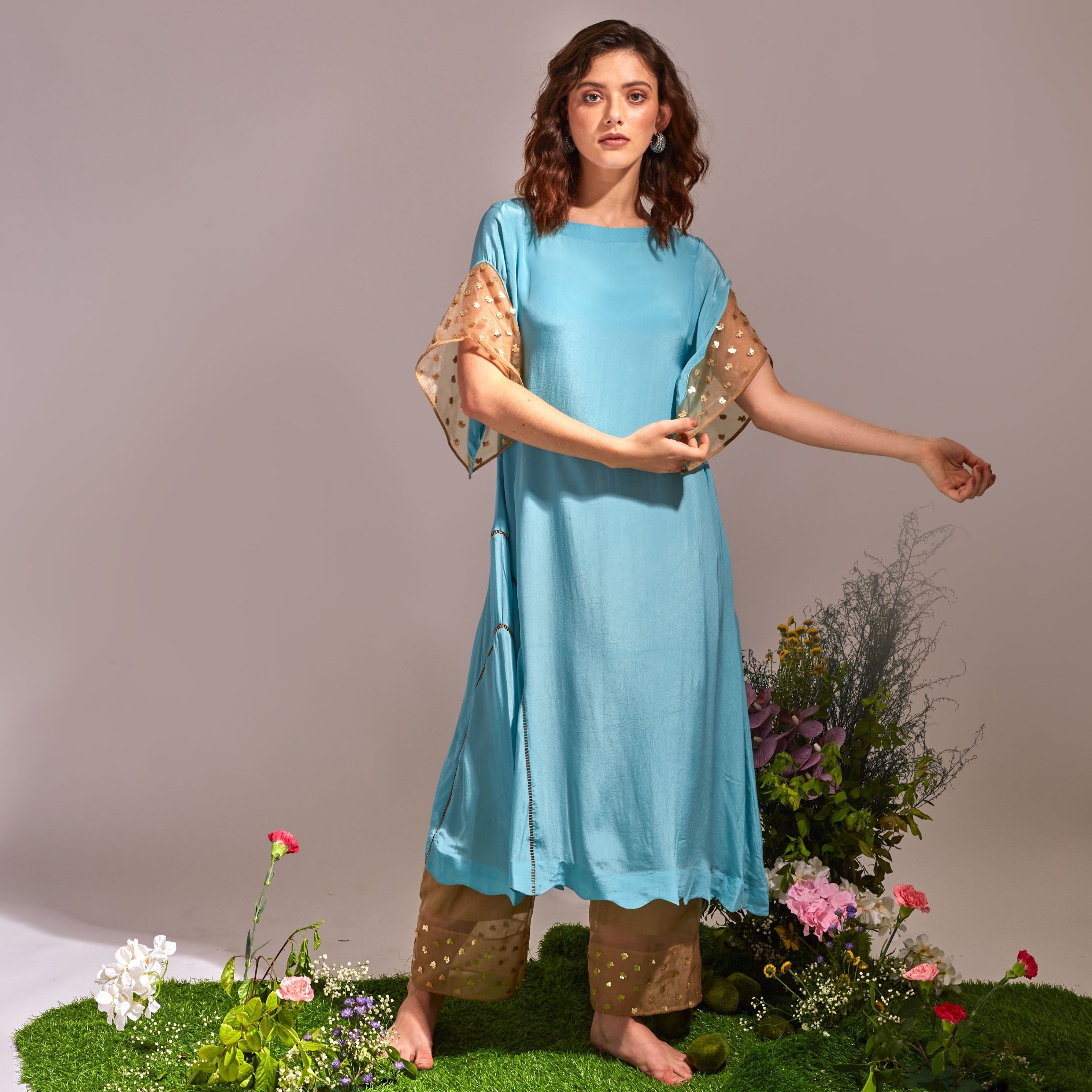 Woman in a blue dress standing on a grassy patch with flowers and plants around her.