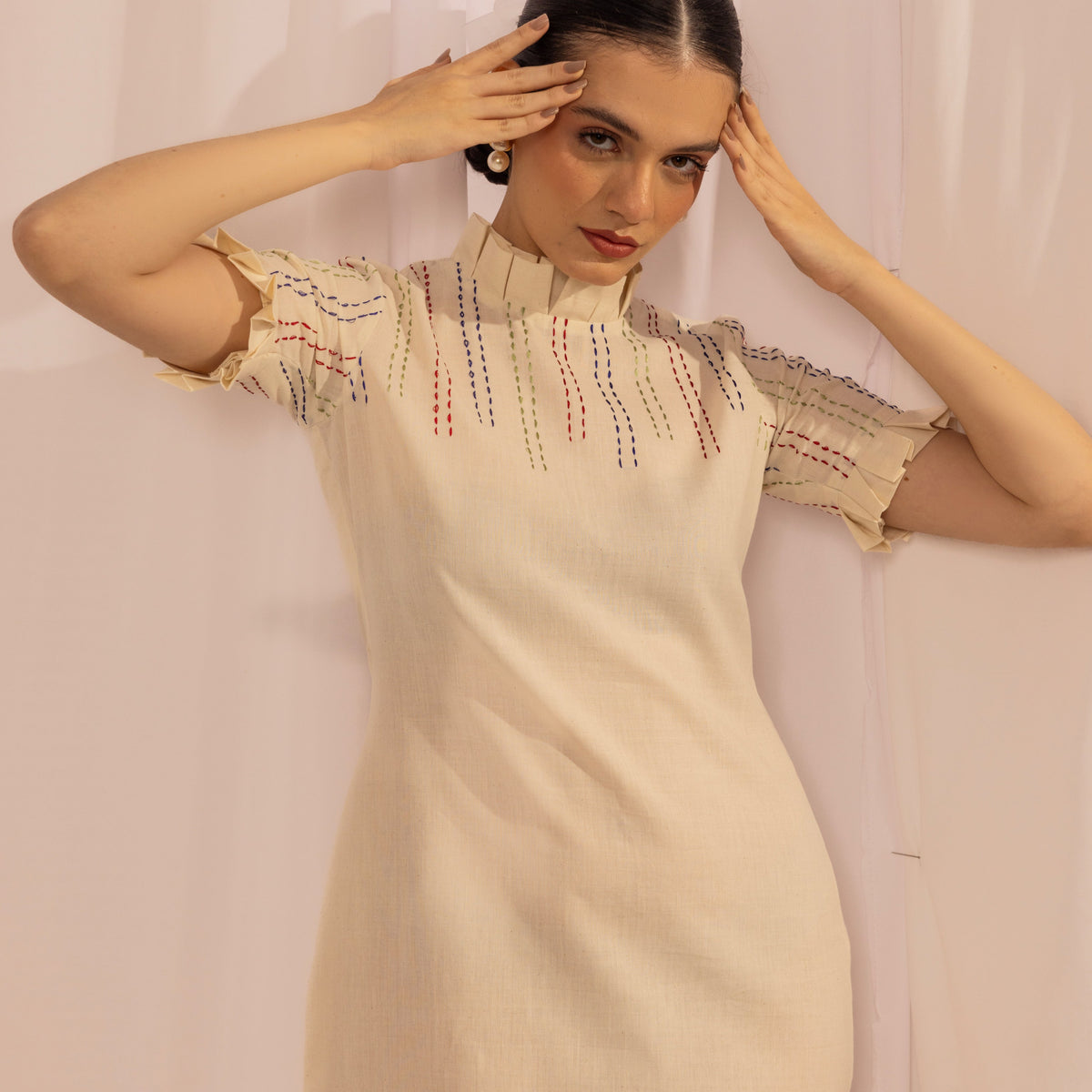 Woman wearing a beige dress with intricate patterns against a white curtain background