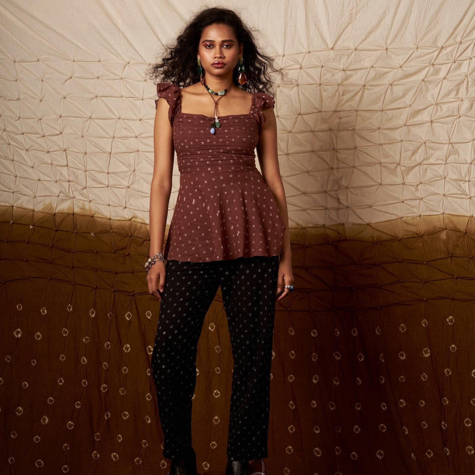 Woman standing on a suitcase wearing a patterned top and pants against a textured wall.