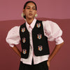 Person wearing a black embroidered vest over a white shirt on a pink background